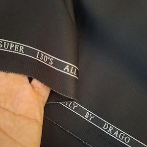 Puede incluir: Primer plano de tela negra con un brillo sutil. La tela tiene una etiqueta blanca que dice "SUPER 130'S ALL ITALY BY DRAGO". El material est&aacute; doblado, revelando su textura y calidad. Ideal para sastrer&iacute;a.