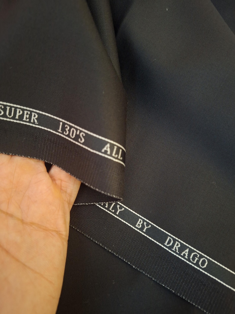 Puede incluir: Primer plano de tela negra con un brillo sutil. La tela tiene una etiqueta blanca que dice "SUPER 130'S ALL ITALY BY DRAGO". El material est&aacute; doblado, revelando su textura y calidad. Ideal para sastrer&iacute;a.