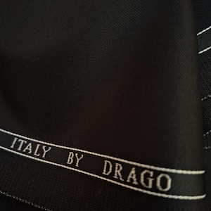 Puede incluir: Primer plano de tela negra con un sutil tejido diagonal. Se ve una cinta blanca con el texto "ITALY BY DRAGO". La tela parece ser un material de alta calidad, posiblemente para trajes o sastrer&iacute;a.