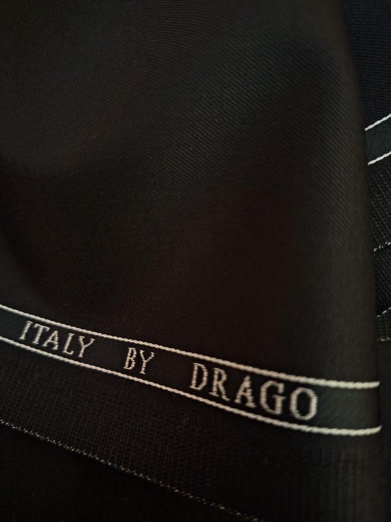 Puede incluir: Primer plano de tela negra con un sutil tejido diagonal. Se ve una cinta blanca con el texto "ITALY BY DRAGO". La tela parece ser un material de alta calidad, posiblemente para trajes o sastrer&iacute;a.