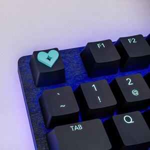 Può includere: Una tastiera meccanica nera con retroilluminazione blu. La tastiera ha un copritasto personalizzato con un design a cuore spezzato in azzurro.
