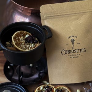May include: A tan paper pouch labelled Chest of Curiosities Est 2023 Apothecary & Witchery, next to a small black cast iron pot containing dried orange slices, berries, and herbs, with additional dried fruit and herbs scattered nearby.
