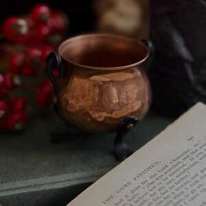 May include: A small, hammered copper cauldron with a rounded body and a wide opening. The vessel features two dark, curved metal handles on the sides and rests on three short, dark metal legs. The surface of the copper has a warm, metallic sheen.