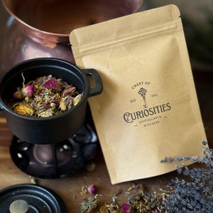 May include: A black cast iron pot filled with dried flowers and herbs, beside a brown paper bag with the text "Chest of Curiosities Apothecary & Witchery". The bag features a key logo and the year 2023. Dried lavender and rose buds are scattered around.