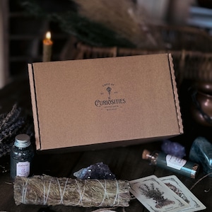 May include: A rectangular brown cardboard box featuring a black logo that reads Chest of Curiosities Apothecary & Witchery with a key icon. The box is surrounded by a small glass jar, a bundle of dried herbs, two illustrated cards, and crystals.