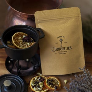 May include: A brown paper pouch labelled Chest of Curiosities Est 2013 Apothecary & Witchery with a key graphic. Next to it is a small black cast iron pot containing dried orange slices, berries, and herbs, with a matching lid and dried lavender.
