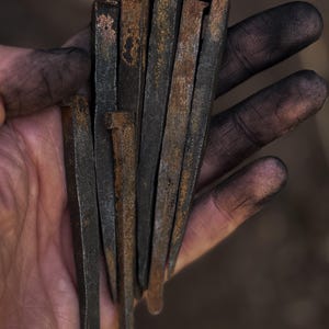 May include: A collection of six hand-forged iron nails with a dark, weathered finish and visible rust patches. Each nail features a square-cut shank and a flat, slightly irregular head, showcasing a raw, industrial texture consistent with traditional blacksmithing techniques.