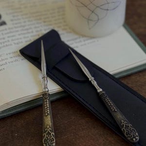 May include: Two silver-toned metal letter openers with pointed tips and ornate, dark-finished handles featuring intricate floral and leaf engravings. The letter openers are positioned on a black rectangular sleeve with a smooth, matte texture.