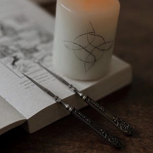 May include: A white pillar candle with a black Celtic knot design is lit and placed next to two silver-toned metal letter openers with ornate, textured handles resting on an open book with visible printed text and a date of 1647.