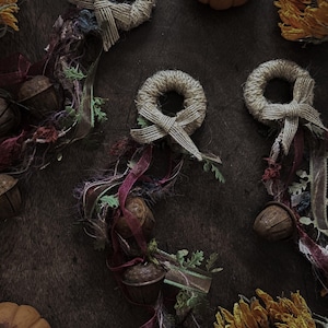 May include: Three decorative ornaments featuring small wreaths wrapped in tan twine with matching bows. Each wreath is adorned with dangling dark brown metal bells, frayed burgundy and purple fabric ribbons, and small green artificial foliage.