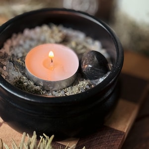 May include: A lit, round, pink candle in a silver holder sits in a black bowl filled with white and green herbs and small stones. A black, polished stone rests beside the candle. The bowl is on a wooden surface.