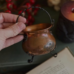 May include: A small, round copper cauldron featuring a hammered texture, a dark metal twisted wire handle, and three curved metal feet. The metallic surface shows natural patina and variations in colour, with a wide opening at the top.