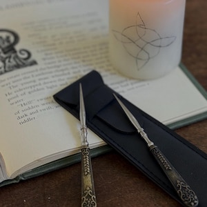 May include: Two silver-toned metal letter openers with ornate, dark-finished handles featuring intricate leaf and scroll patterns. The letter openers are resting on a black rectangular case, positioned next to an open book and a lit pillar candle.