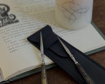 Puede incluir: Dos abrecartas de metal plateado con mangos oscuros decorados con intrincados patrones de hojas y volutas. Los abrecartas descansan sobre un estuche rectangular negro, junto a un libro abierto y una vela de pilar encendida.