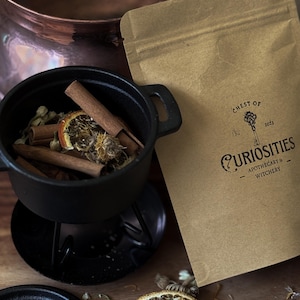 May include: A black cast iron cauldron filled with cinnamon sticks and dried botanicals, positioned next to a tan paper pouch labelled Chest of Curiosities Apothecary & Witchery 2023. A dried orange slice and loose herbs are scattered nearby.