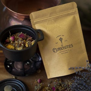 May include: A brown paper pouch labelled Chest of Curiosities Est 2023 Apothecary & Witchery with a key graphic, positioned next to a small black cast iron pot filled with a blend of dried flower buds, yellow petals, and green herbs.