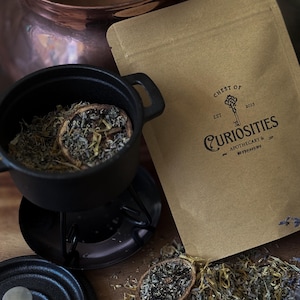 May include: A tan paper pouch labelled Chest of Curiosities Apothecary & Witchery Est 2023 with a key graphic. Next to it is a small black cast iron pot containing dried herbs and citrus slices, with a matching lid and a copper vessel nearby.
