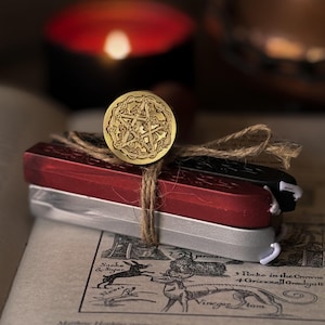 May include: A gold-coloured wax seal stamp featuring a pentagram design, tied with twine to three rectangular wax sticks in red, black, and silver. The wax sticks are stacked horizontally and secured together with a natural fibre string.