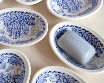 May include: Oval-shaped white and blue soap dishes with a floral design. One dish holds a light blue bar of soap with the text "Crabtree & Evelyn". The dishes have a detailed blue pattern around the rim and inside.