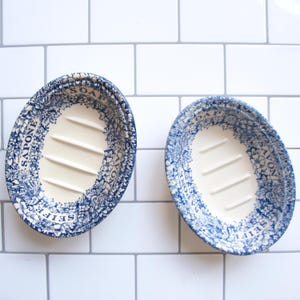May include: Two oval-shaped ceramic soap dishes featuring a cream-colored center with raised ridges and a blue and white floral pattern around the rim. The word SOAP is printed on the top, with SAPONE, SAVON, and SEIFE on the sides.