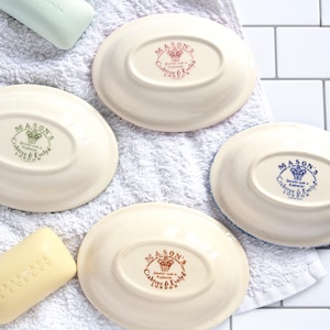May include: Four oval-shaped cream-colored ceramic soap dishes, each featuring a central stamp with a crown logo and the text Mason's, Crabtree & Evelyn London. Two bars of soap are included, one pale green and one pale yellow with embossed text.