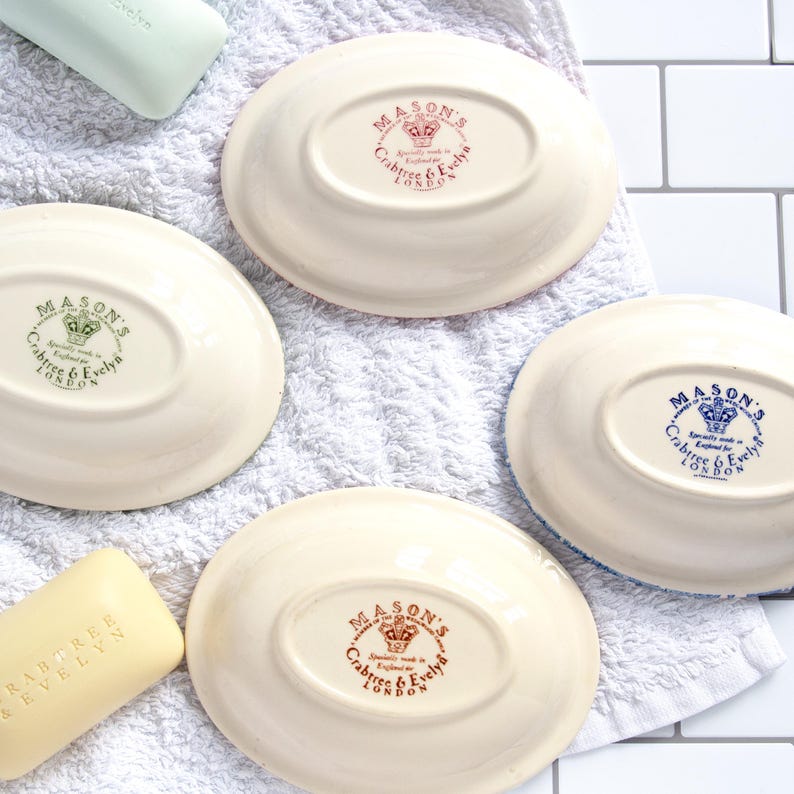 May include: Four oval-shaped cream-colored ceramic soap dishes, each featuring a central stamp with a crown logo and the text Mason's, Crabtree & Evelyn London. Two bars of soap are included, one pale green and one pale yellow with embossed text.