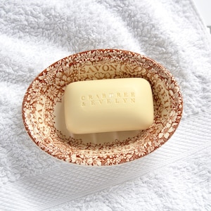 May include: A rectangular bar of cream-colored Crabtree & Evelyn soap resting in an oval ceramic dish. The dish features a brown and white floral pattern with the words SAVON, SOAP, SEIFE, and SAPONE embossed around the inner rim.