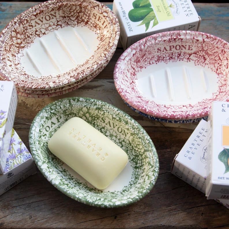 May include: Three oval ceramic soap dishes with floral patterns in brown, red, and green, each featuring the word soap in different languages. A rectangular bar of cream-colored soap is placed inside the green dish, surrounded by boxed soap products.