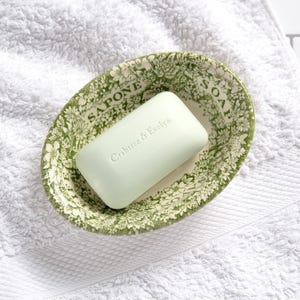May include: A rectangular light green bar of soap with the embossed text Crabtree & Evelyn, resting inside an oval ceramic dish featuring a green and white floral pattern and the words SAPONE and SOAP printed along the inner rim.