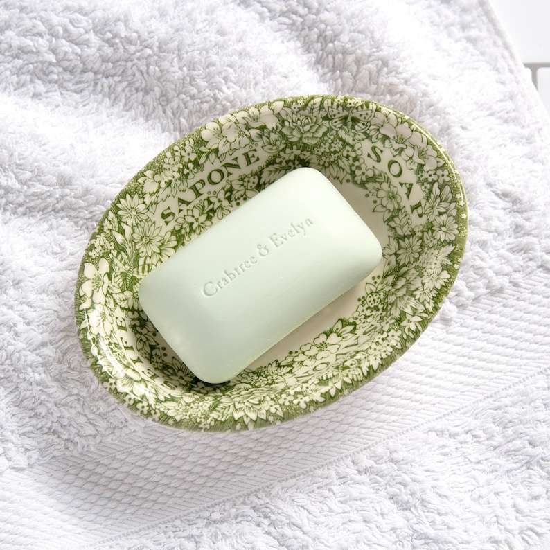 May include: A rectangular light green bar of soap with the embossed text Crabtree & Evelyn, resting inside an oval ceramic dish featuring a green and white floral pattern and the words SAPONE and SOAP printed along the inner rim.