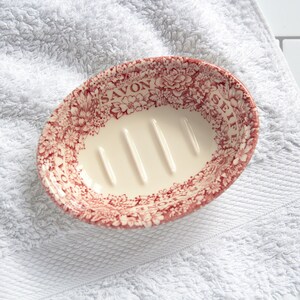 May include: An oval ceramic soap dish featuring a cream-colored interior with four raised drainage ridges. The rim is decorated with a red floral pattern and the words SOAP, SAVON, and SEIFE printed in a serif font around the edge of the dish.