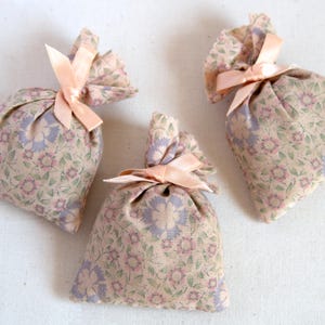 May include: Three floral fabric sachets tied with peach-coloured ribbons. The sachets feature a beige background with a pattern of pink and purple flowers and green leaves. The ribbons are tied in bows.