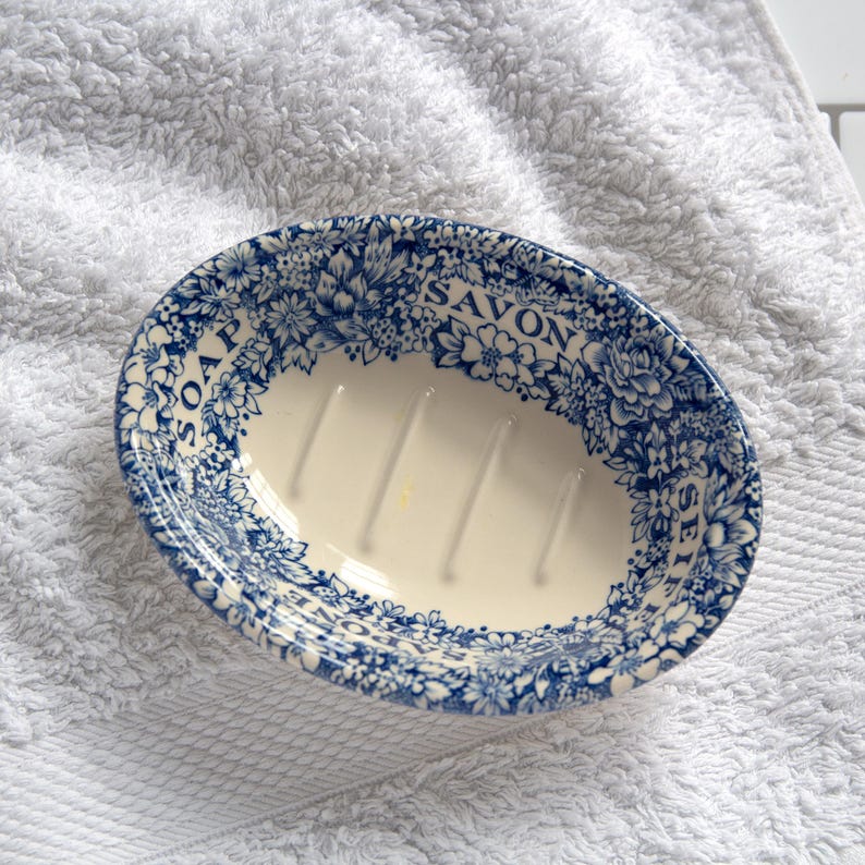 May include: An oval ceramic soap dish with a cream-colored interior featuring three raised drainage ridges. The rim is decorated with a blue and white floral pattern and the printed text "SOAP", "SAVON", and "SEIFE" around the border.