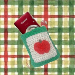 May include: A red Canon camera partially inserted into a handmade green and white crochet case. The case features a red apple design. The background is a plaid pattern in red, green, and white.