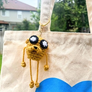 May include: A handmade crochet keychain featuring a yellow dog-like character with black and white eyes, hanging from a gold-colored ring. The keychain is attached to a beige tote bag with a blue heart and the word "CONFIDENCE" printed on it.