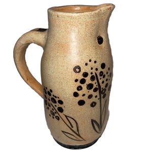 May include: A beige ceramic pitcher with a handle and spout. The pitcher is decorated with black floral designs. The pitcher has a speckled texture and a dark brown base.