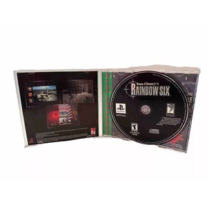 May include: A PlayStation game case for Tom Clancy's Rainbow Six. The case is open and the disc is visible. The disc is black with a green and white target design. The text "Tom Clancy's Rainbow Six" is printed in white on the disc.