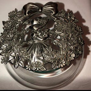 May include: Silver-toned metal lid with a floral design and a girl figurine in the center. The lid is designed to fit a glass jar.