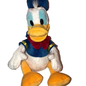May include: A plush Donald Duck toy. The toy features a blue sailor hat, a blue sailor shirt with gold trim, a red bow tie, and yellow feet and beak. The toy has white feathers and blue eyes.