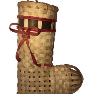May include: A woven basket shaped like a boot, featuring a handle and a decorative red ribbon bow. The basket is made of light brown woven material, with a darker brown band around the top. The boot shape is approximately 10 inches tall.