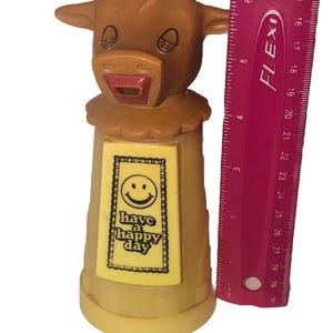May include: A brown cow-shaped figurine with a smiling face and the words "have a happy day" on a yellow label. The figurine is approximately 5 inches tall. A pink ruler is next to the figurine.