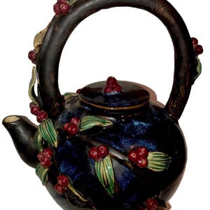 May include: A dark blue ceramic teapot with a large handle and a lid. The teapot is decorated with red berries and green leaves. The handle and spout are dark brown. The teapot is likely used for brewing tea.