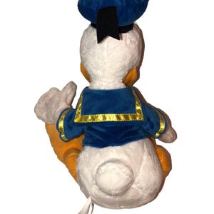 May include: A plush Donald Duck toy, viewed from the back, with a white body, orange feet, and a blue sailor suit. The hat is blue with a black ribbon. The toy is soft and cuddly, perfect for Disney fans.