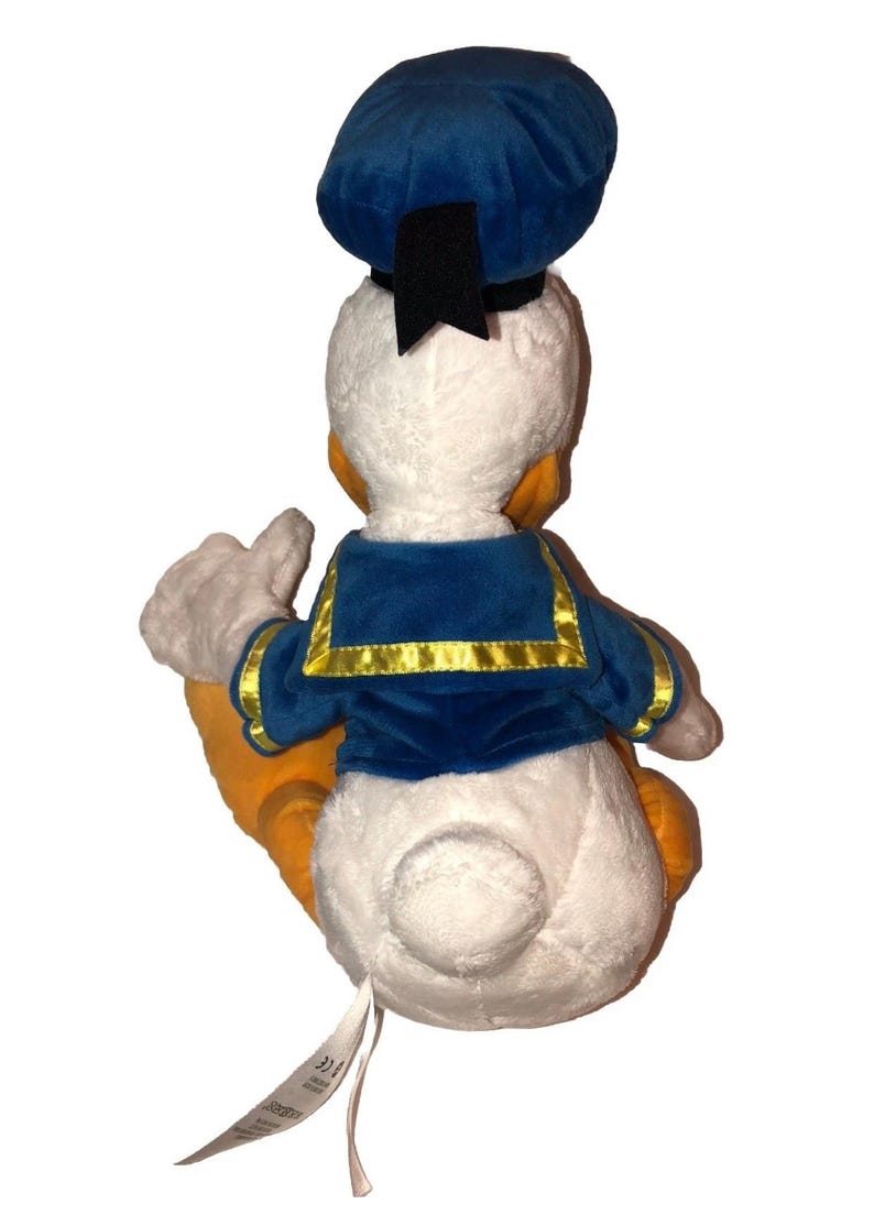 May include: A plush Donald Duck toy, viewed from the back, with a white body, orange feet, and a blue sailor suit. The hat is blue with a black ribbon. The toy is soft and cuddly, perfect for Disney fans.