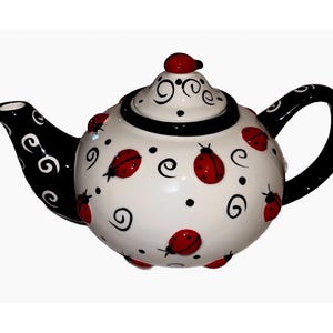 May include: A ceramic teapot with a white base, adorned with red ladybug designs and black swirls. The spout and handle are black, and the lid features a red ladybug. A decorative kitchenware item.