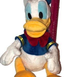 May include: A plush Donald Duck toy, featuring a white body, blue sailor shirt, and a red bow tie. The duck has a yellow beak and feet, with a blue hat. A ruler is placed next to the toy.
