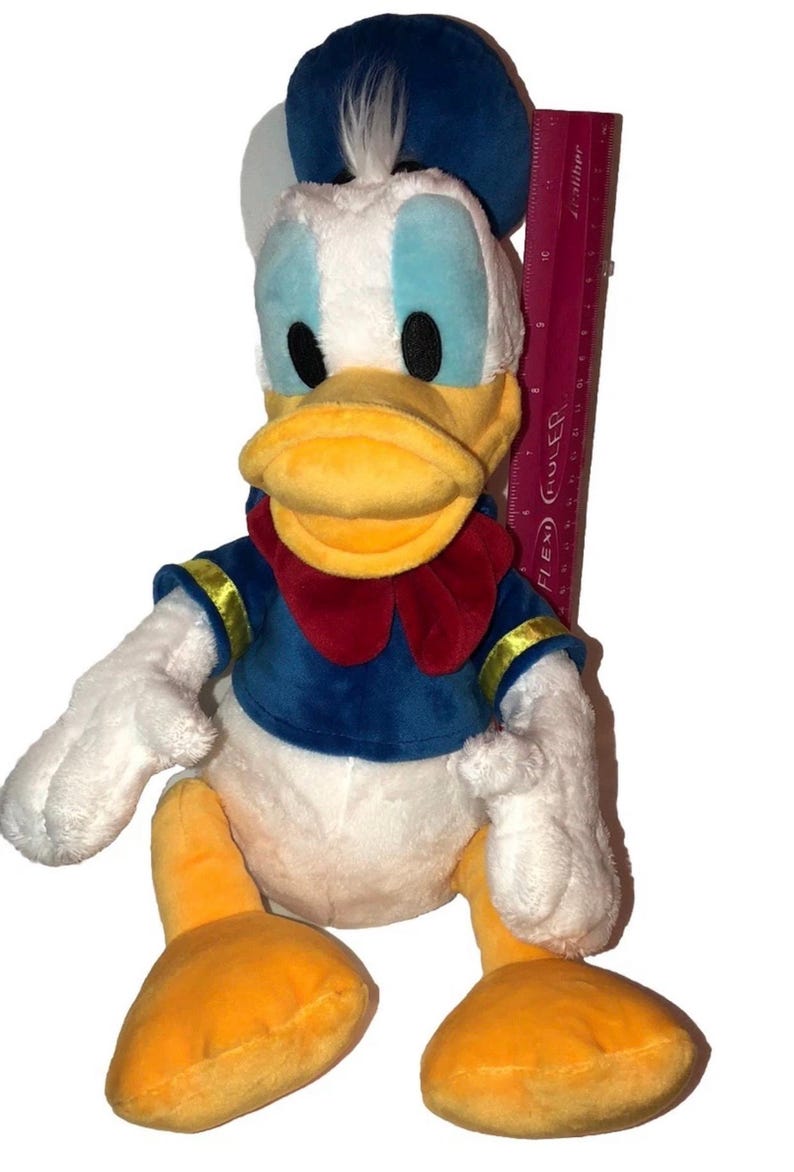 May include: A plush Donald Duck toy, featuring a white body, blue sailor shirt, and a red bow tie. The duck has a yellow beak and feet, with a blue hat. A ruler is placed next to the toy.
