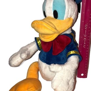 May include: A plush Donald Duck toy, featuring a white body, blue sailor shirt with gold trim, and a red bow tie. The duck has a yellow beak and feet, and a blue sailor hat. A ruler is next to the toy.