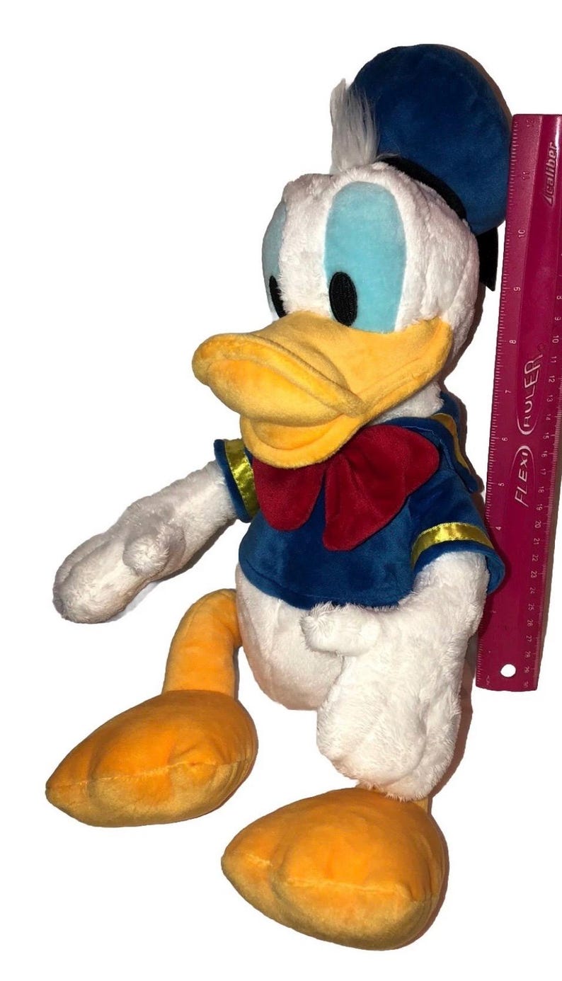 May include: A plush Donald Duck toy, featuring a white body, blue sailor shirt with gold trim, and a red bow tie. The duck has a yellow beak and feet, and a blue sailor hat. A ruler is next to the toy.