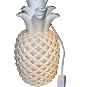 May include: White pineapple-shaped wall sconce with a textured surface and a light inside. The lamp has a switch and a white cord. The pineapple design features a crown of leaves at the top.